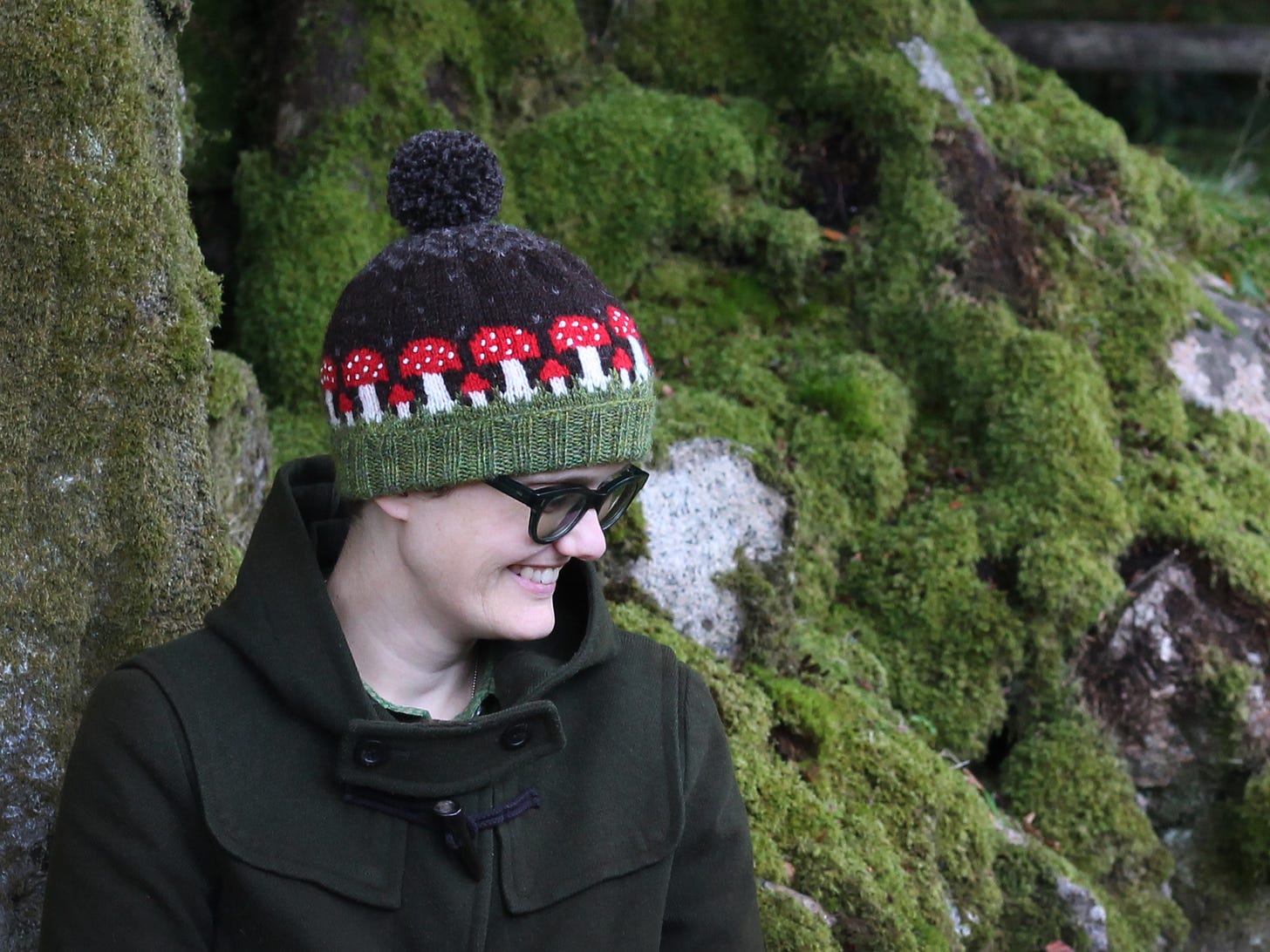 Image decription: a headshot of Katie, a white person with glasses looking sideways and down to the right of frame. Katie is wearing a dark green duffle coat and a woolly bobble hat with green brim, dark brown background and a ring of joyous red and white mushrooms all around. 