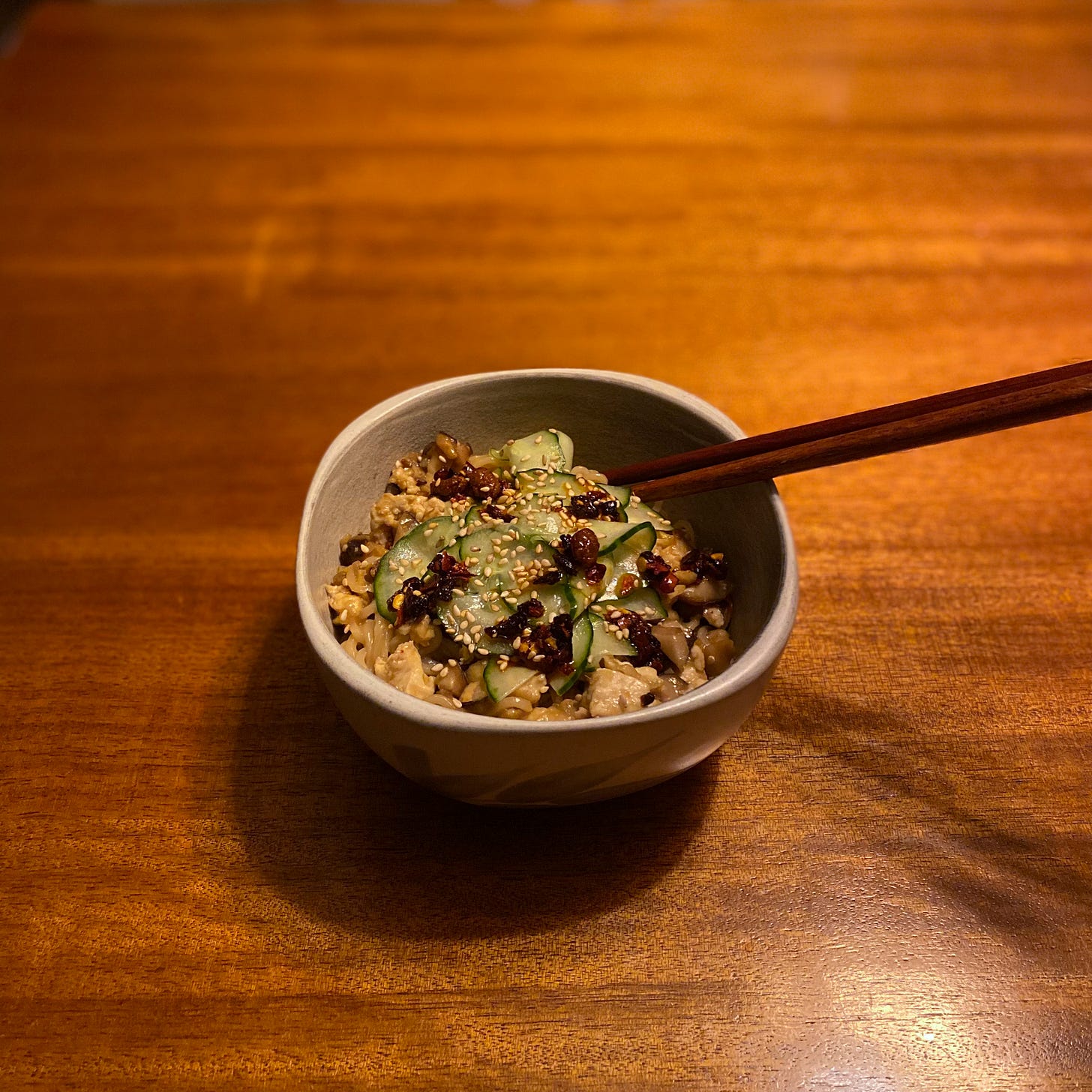 An off-white bowl of the tofu & shiitake noodle stir fry, with brown chopsticks sticking out at the side. On top are thin slices of quick pickled cucumber, sesame seeds, and chili crisp.