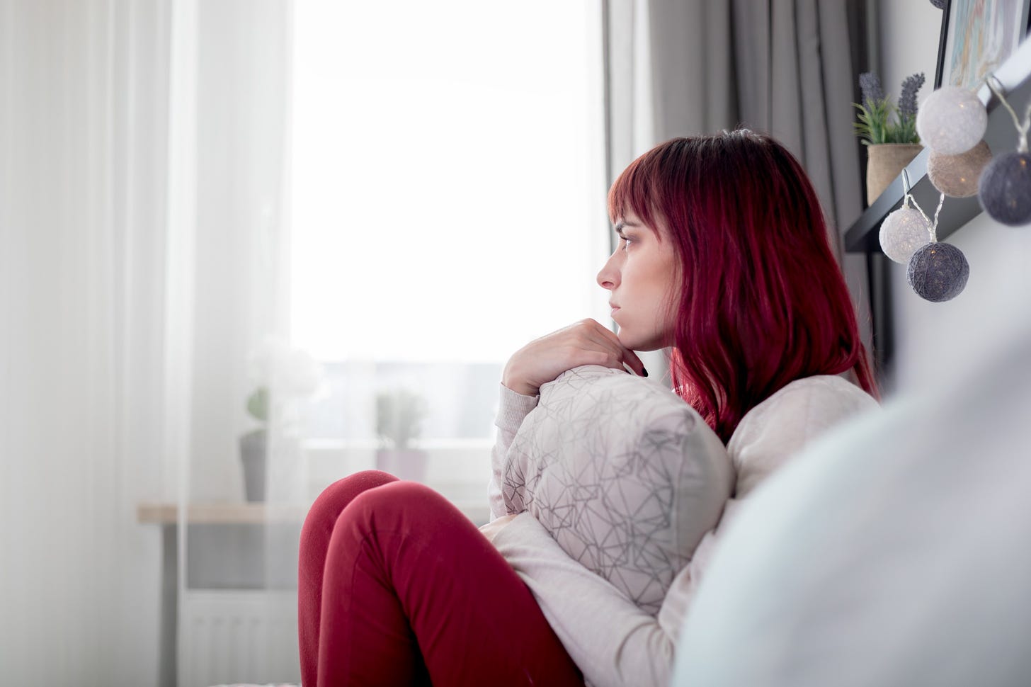 A pensive woman with bright red hair stares out of the window and hugs a pillow.
