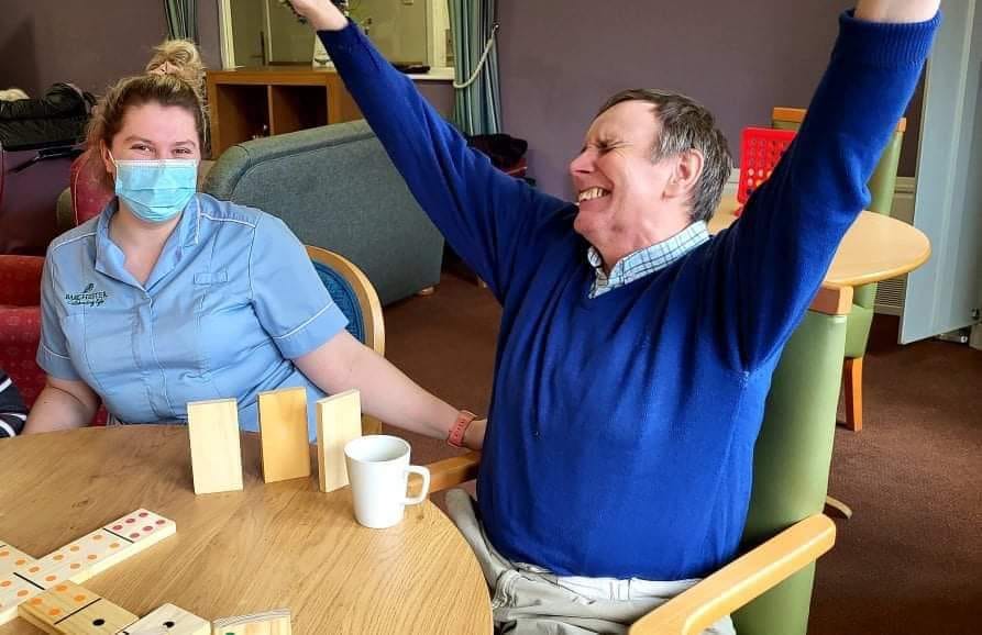 My dad, wearing a blue jumper, celebrating a win during a dominoes match whilst his carer sits next to him.