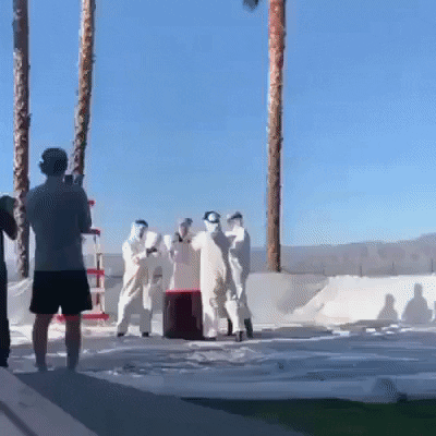 Four men in white hazmat type suits drump liquid into a large white drum and run away as red foam erupts about seven feet high
