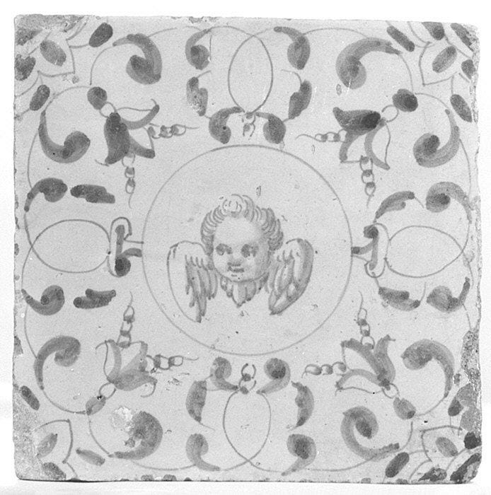 Ceramic tile with face and wings at center, floral decoration surrounding.