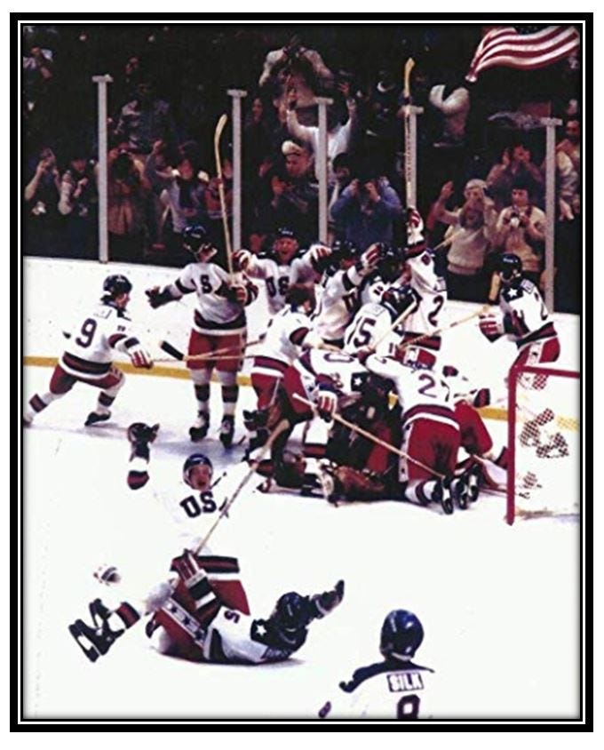 Classic picture of the US hockey team at the moment they win.  Players are piled on top of each other, celebrating. The crowd behind them waves flags and cheers.