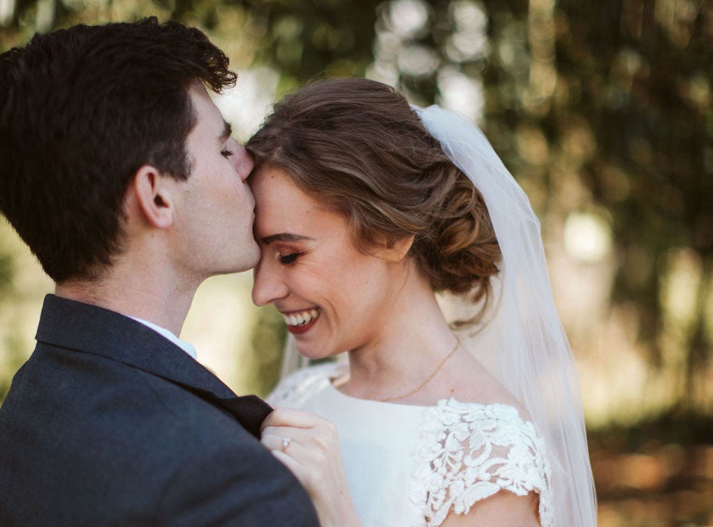 A dark-haired man kissing his smiling bride on the forehead, with dappled sunlight in the background