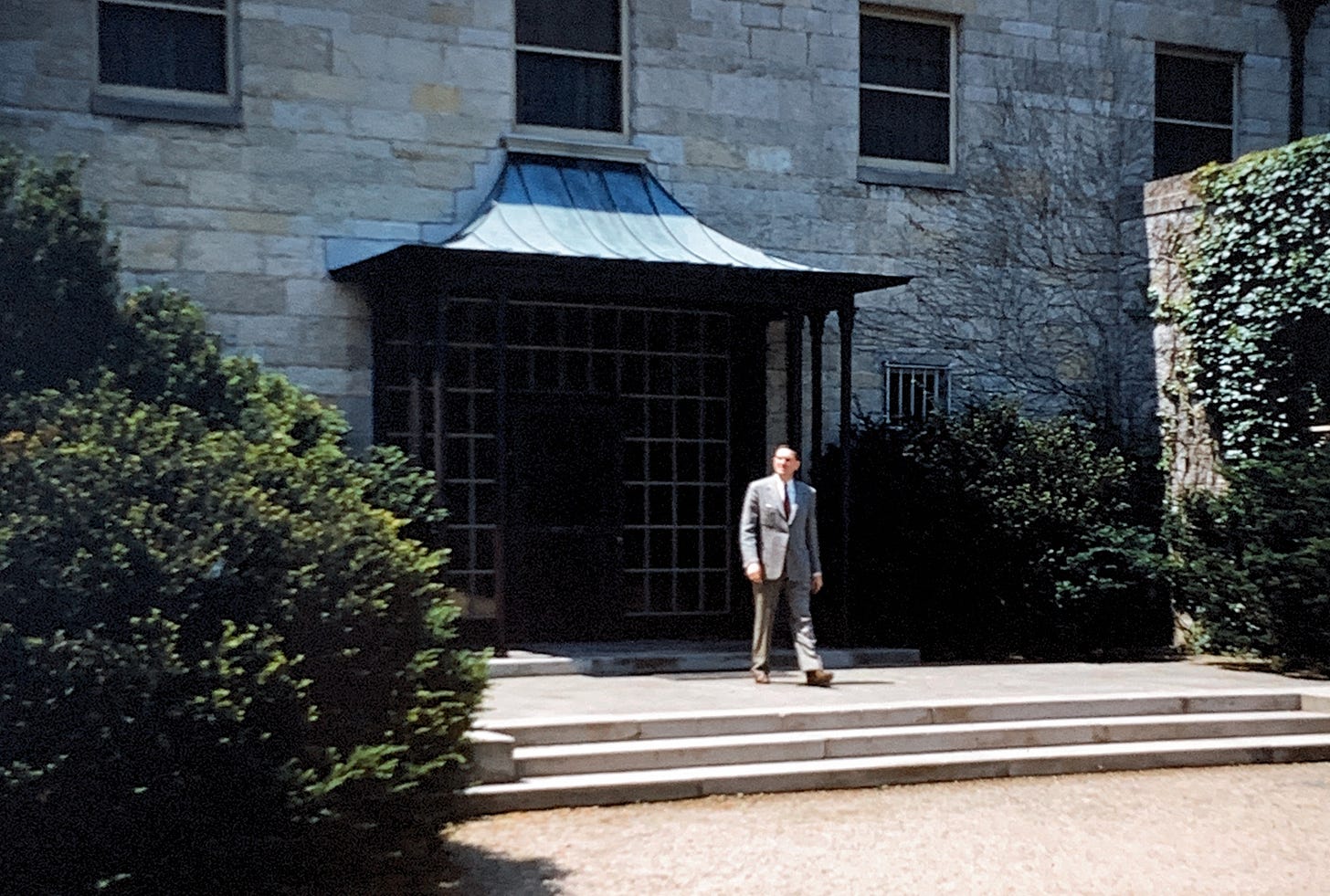 A man in a grey suit walking out of a white stone building and down three stone steps.