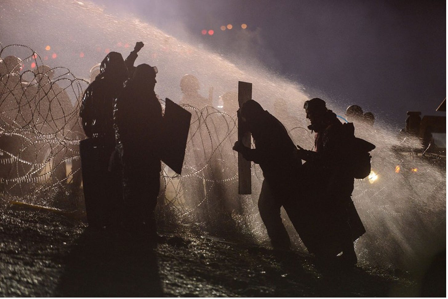 A picture of water protectors at Standing Rock being sprayed with water by police from across a barbed wire fence. The barbed wire has small icicles hanging from it.