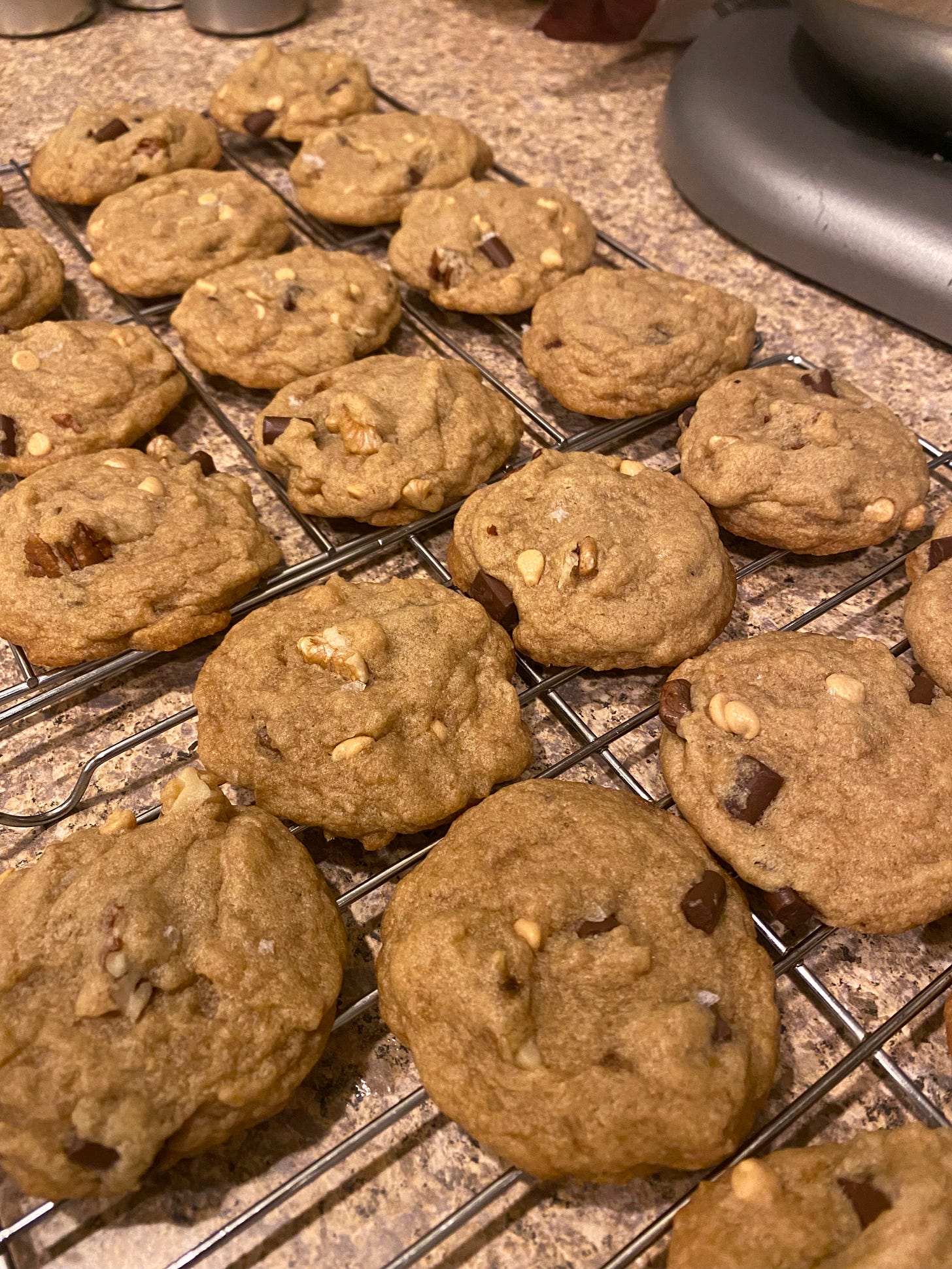 On a cooling rack, a batch of chocolate chunk cookies with nuts and peanut butter chips. Flakes of salt are visible on some of them.