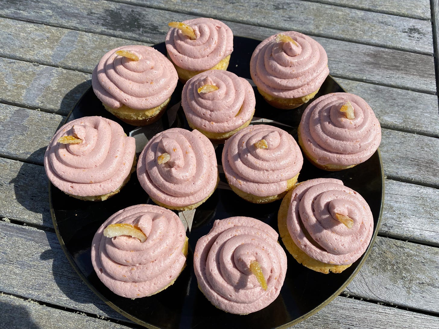 A plate of cupcakes on a wooden picnic table. They are frosted with pink buttercream pipped in a spiral pattern, and each one is topped with a small piece of candied lemon.