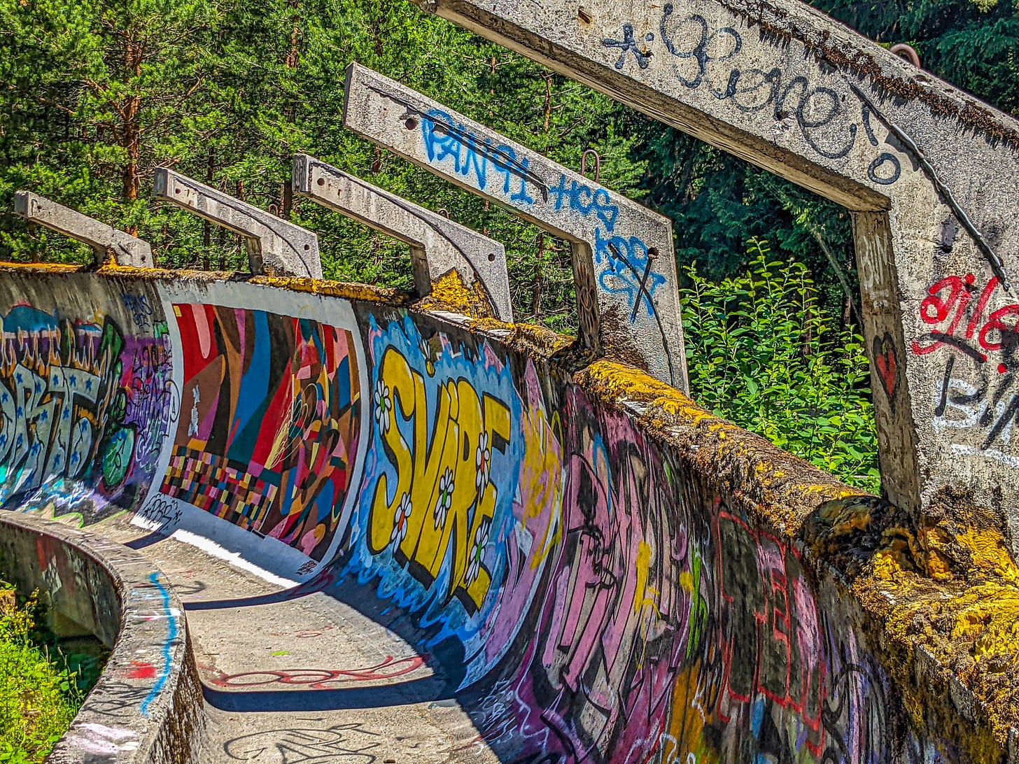 One of the runs curving away, the concrete painted with different murals including the word "SVIRE" in bright yellow next to a geometric painting in reds and blues. The forest looms behind. 
