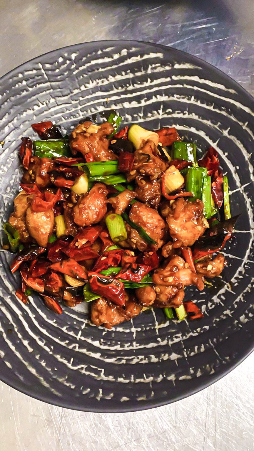 An overhead shot of a dark textured plate in the middle of which is a pile of bite-sized pieces of cooked chicken, spring onions and red chillies.