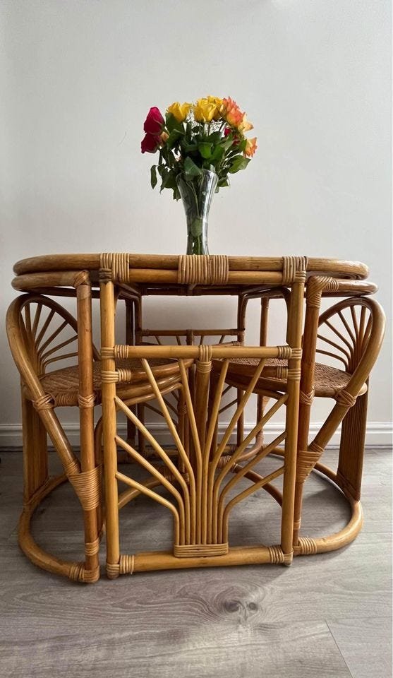Inside an apartment, a table made from cane and bamboo is pushed against the wall. Two matching bamboo chairs are tucked beneath the table's surface. On the table, there is a vase of fresh cut flowers.