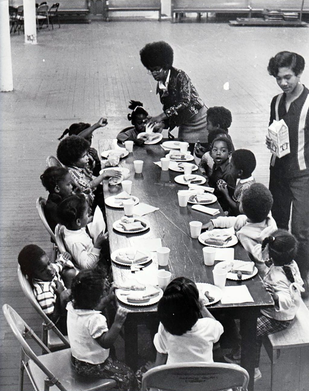 a group of Black children sit at a table eating free breakfast. a Black woman helps a young girl pick up her plate.