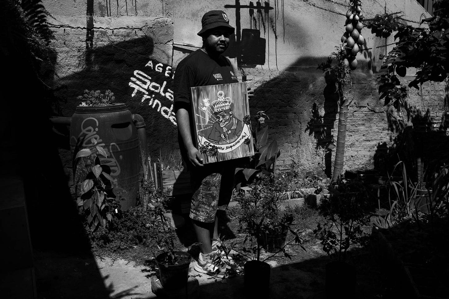 Foto em preto e branco mostra Thiago Vinicius em pé no quintal da agência Solano Trindade segurando uma pintura que mostra um homem com coroa