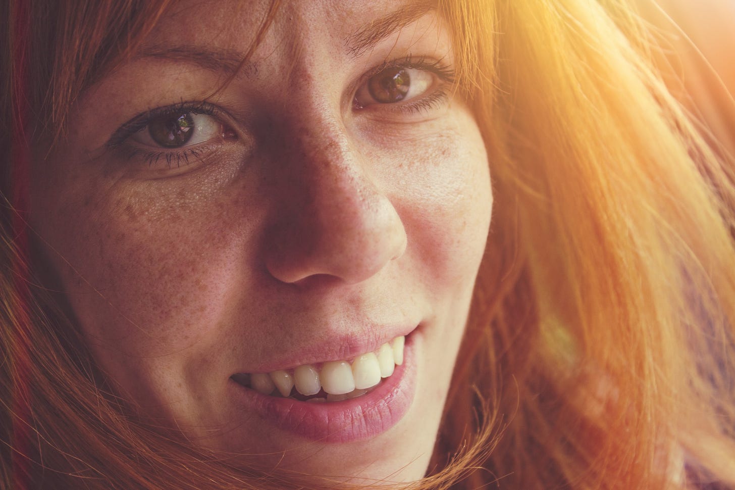 A woman with red hair and brown eyes smiles closely into the camera.