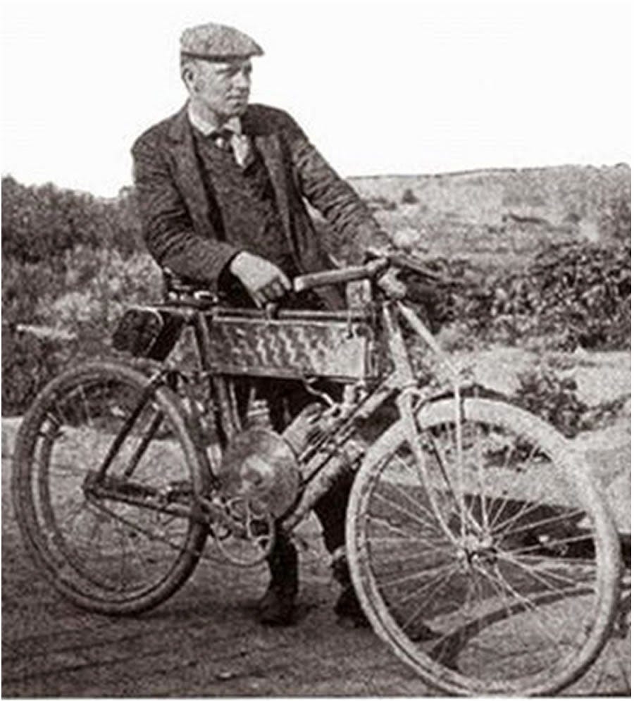 Grainy black and white photo of George A. Wyman and his California motor bicycle. The bike is angled so that the front wheel sits in the bottom right corner and the rear wheel nearly touches the left edge but is raised from the bottom of the photo. The bike bears a closer resemblance to a bicycle, with a double-diamond frame, pedals, crank, and chain and bicyle style handlebars, than a motorcycle. The bits that make it a motorcycle include a metallic tank hanging beneath the top tube, along with a small displacement motor (~200cc), large, exposed metal flywheel, and a leather belt connecting the motor to the rear wheel. Wyman stands to the left of the bike, his right elbow leaning against the seat as his left hand holda the left handlebar. He is wearing a three piece suit, cap, and tie, and looks steadily into the distance. Behind him are bushes and what appears to be an escarpment.