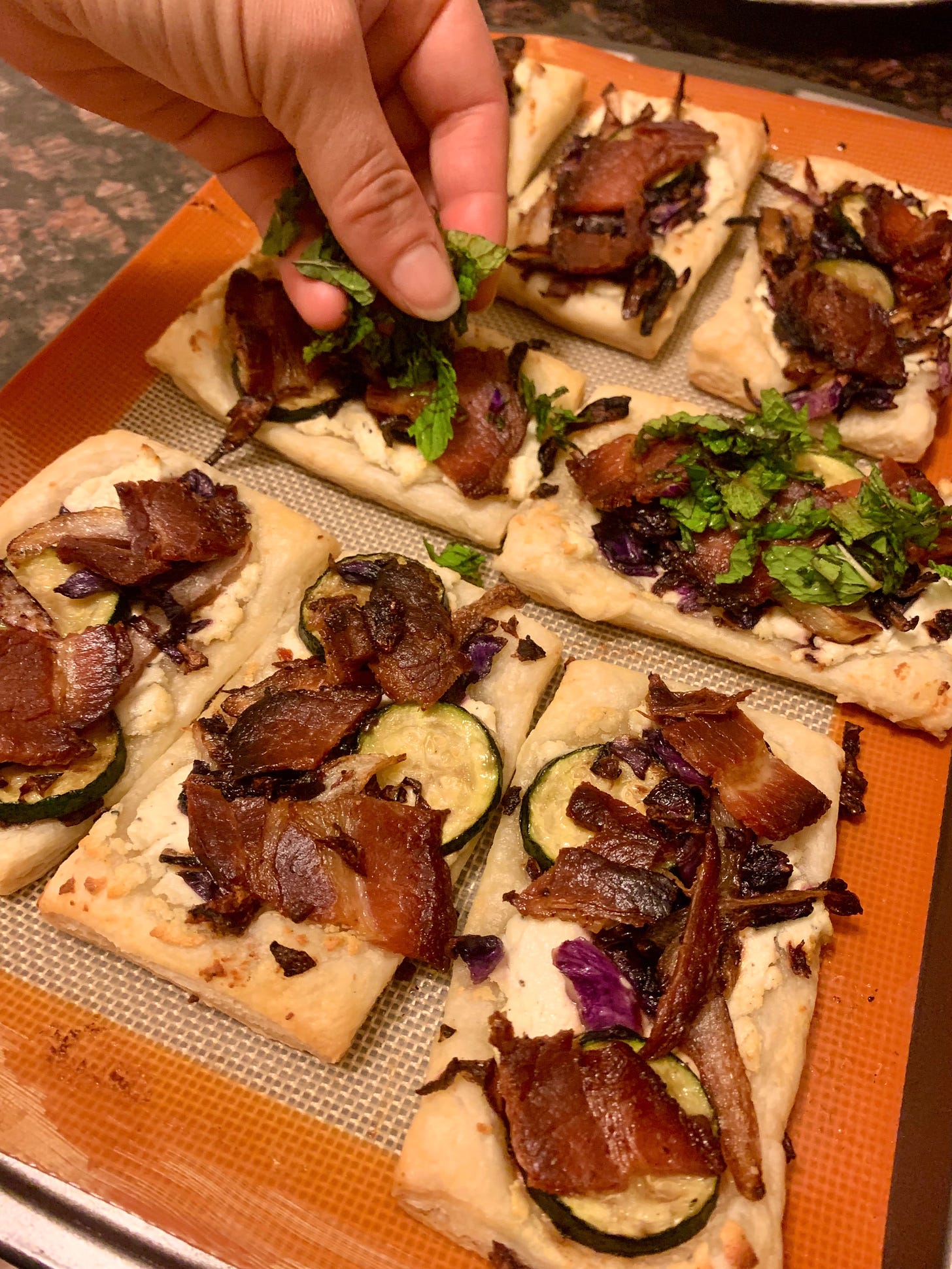 Top view of hand holding mint as it is sprinkled on slices of puff pastry topped with vegetables and bacon