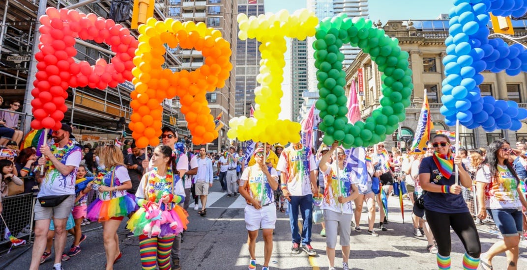 A photo from Toronto Pride, feature a large crowd walking down Yonge Street in Toronto, and 5 people holding large coloured balloon letters that spell PRIDE.