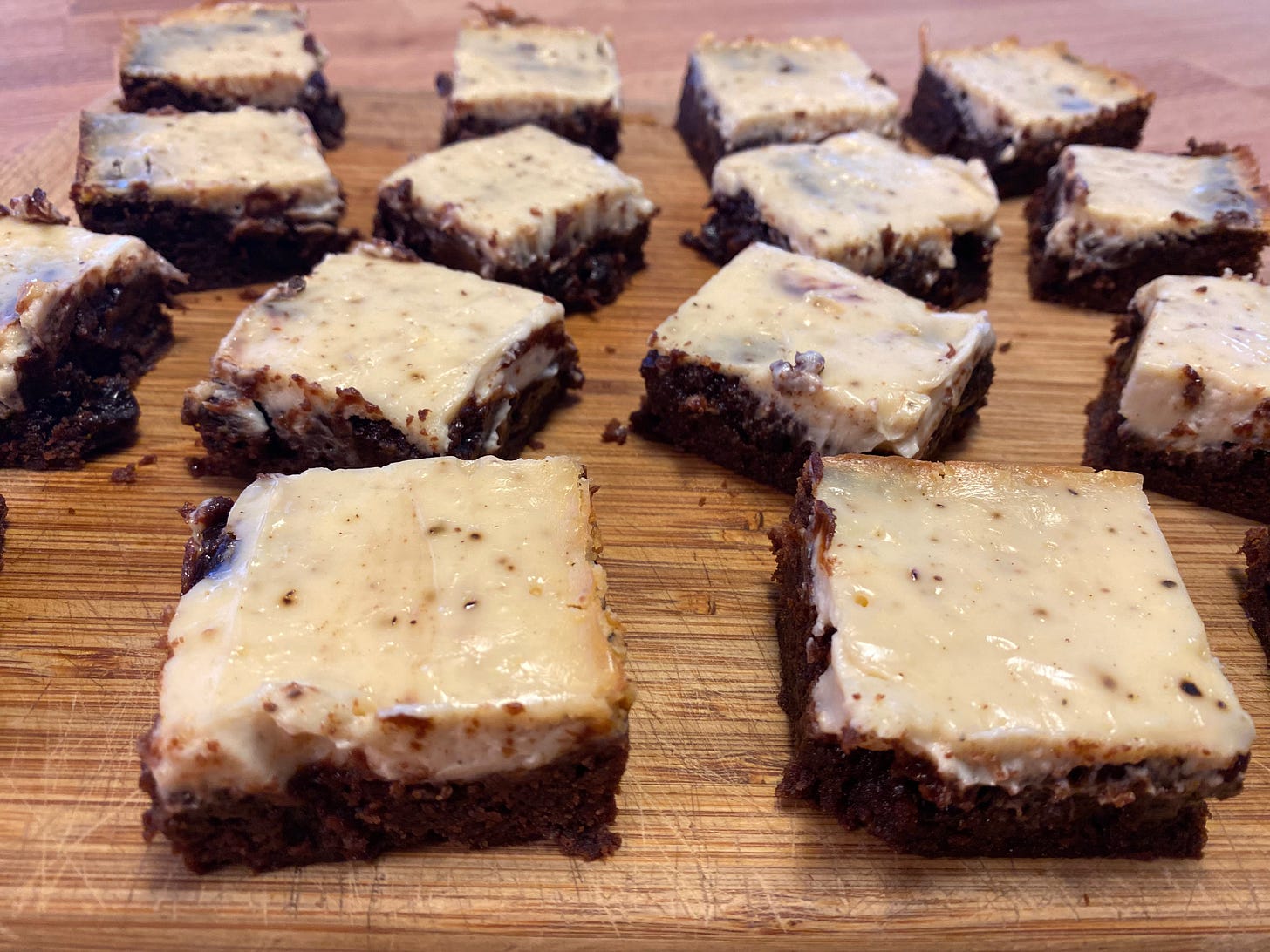 Several rows of brownies arranged on a wooden cutting board. They are cut into squares, and each one is topped with a shiny, creamy cheese layer.