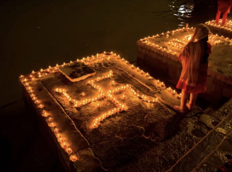 A Dharmic woman dressed in traditional clothes stands before a design of lamps arranged in the shape of a swastika symbol. She is on a river bank at night looking out towards the river while the lamps provide golden light that reflects off the water. There is another swastika lamp design next to her and another beyond that with another person standing by it.