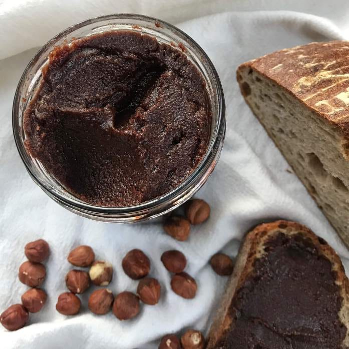 a jar of hazelnut chocolate spread with some sourdough bread in the background and a handful of hazelnuts