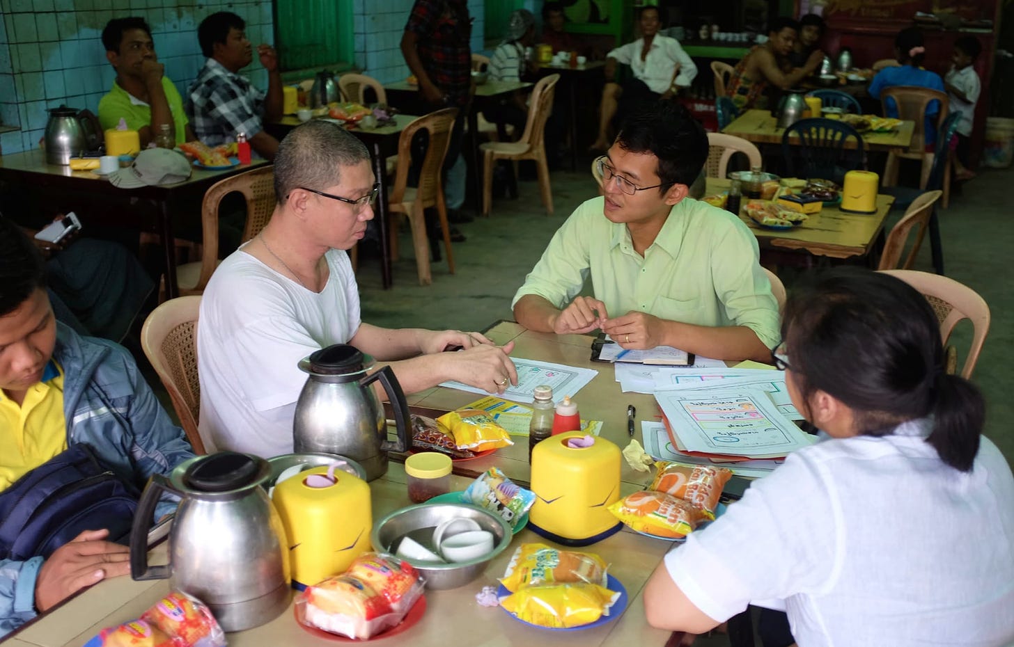 Four people sit at a table featuring packaged snacks, bottles, coffee pots, and letter-size papers, in an informal restaurant. One man, wearing a light-green shirt, is speaking with another man, in a white t-shirt, who is pointing to a paper in front of him.  Two other people are in the foreground, one looking down and one with her back turned.