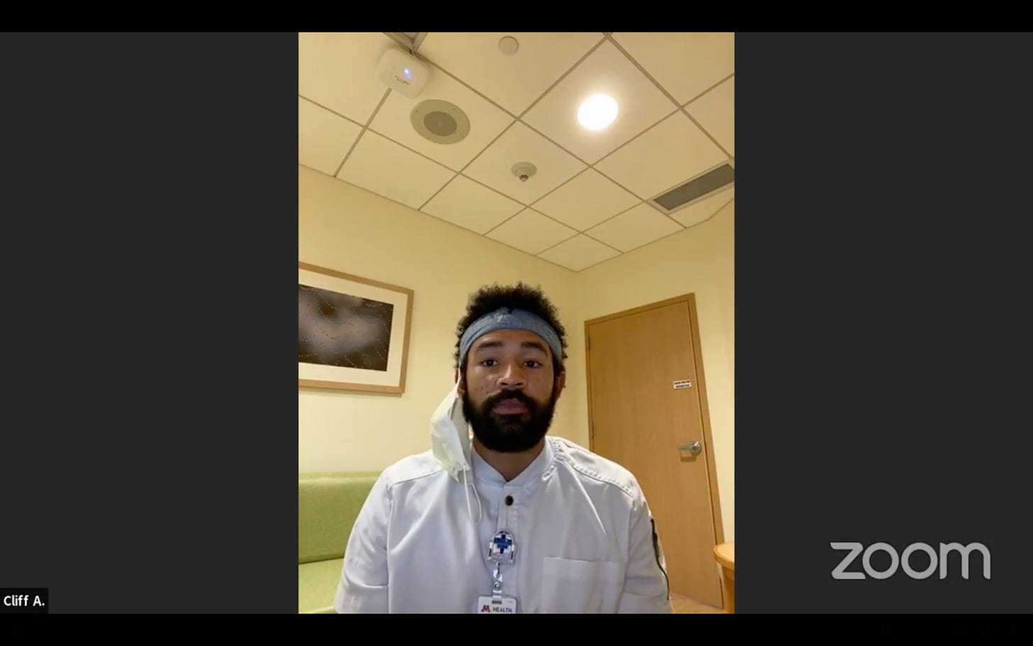 a black man sits in a hospital room on a zoom livestream with a white mask hanging from his ear, in white scrubs