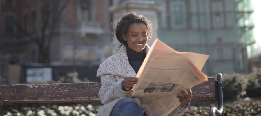 Young woman sitting on bench outside reading newspaper