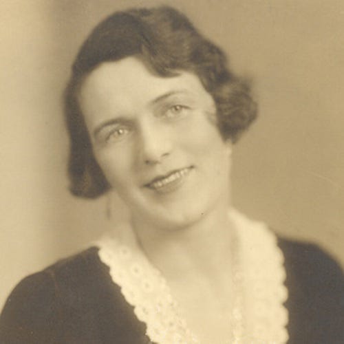 Sepia toned photo of a white woman with dark hair, swept back. She smiles and shows her teeth and looks off to the right shoulder of the photographer