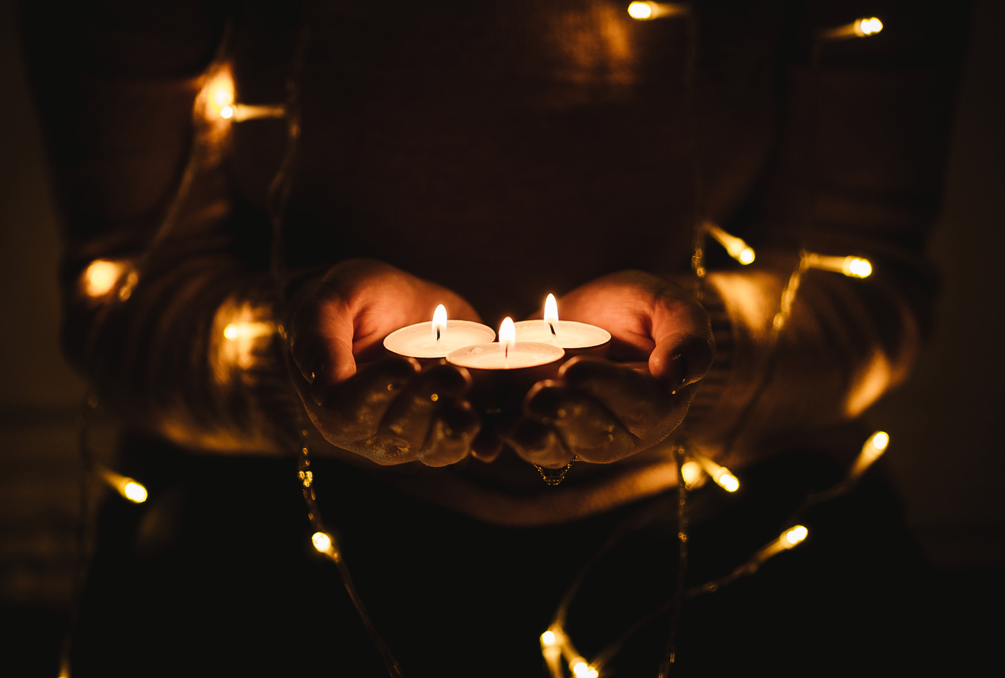 Two outstretched hands holding three votive candles surrounded by small white lights