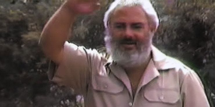 Bert Montoya is pictured from the chest up waving to the camera with his left hand. He is dressed in a beige button-up shirt with short sleeves. He has deep, olive-toned skin and salt-and-pepper colored hair and grown-out beard. He is standing in front of green trees. 