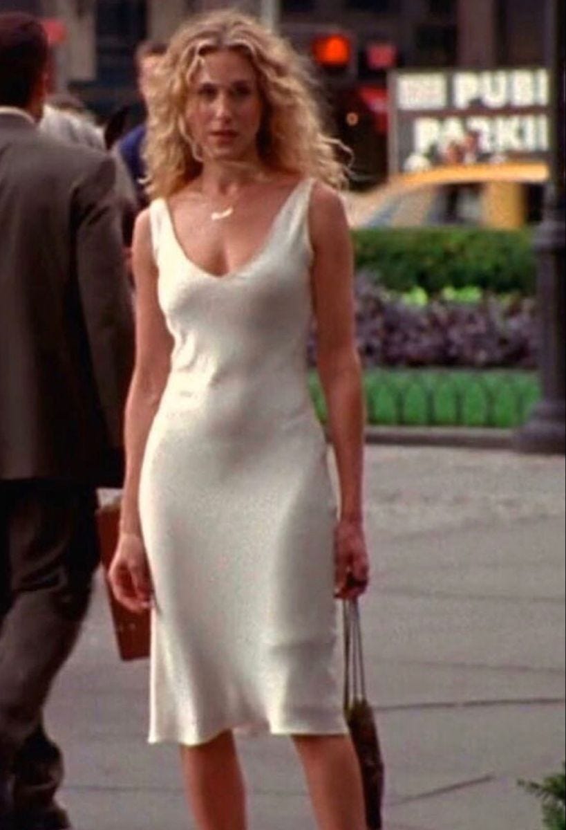 Carrie Bradshaw stands in a white satin slip dress. She's looking sideways at something we can't see but her devastation and shock are obvious. Her hair blows open and free around her face. The dress drapes, clinging because of the wind. She clutches a bag in one hand, both arms hanging by her side as she stands on the street.