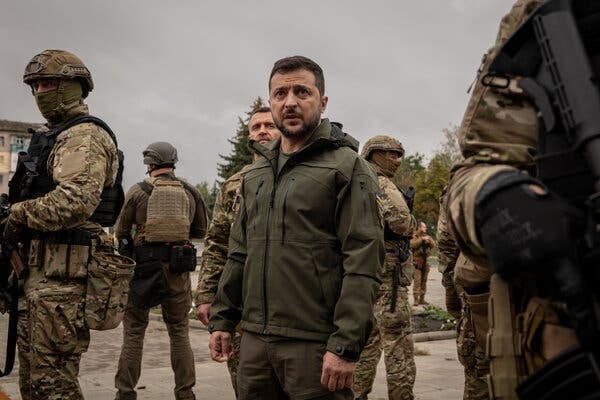 Volodymyr Zelensky, wearing an olive jacket and pants, surrounded by Ukrainian soldiers guarding him. 