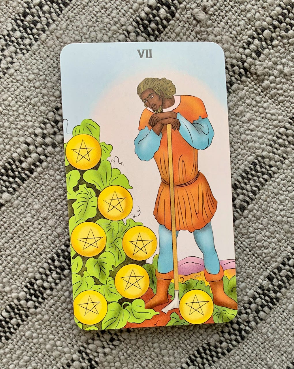 A black man in an orange and blue tunic leans on a hoe and looks at a patch of pumpkins, but the pumpkins are pentacles