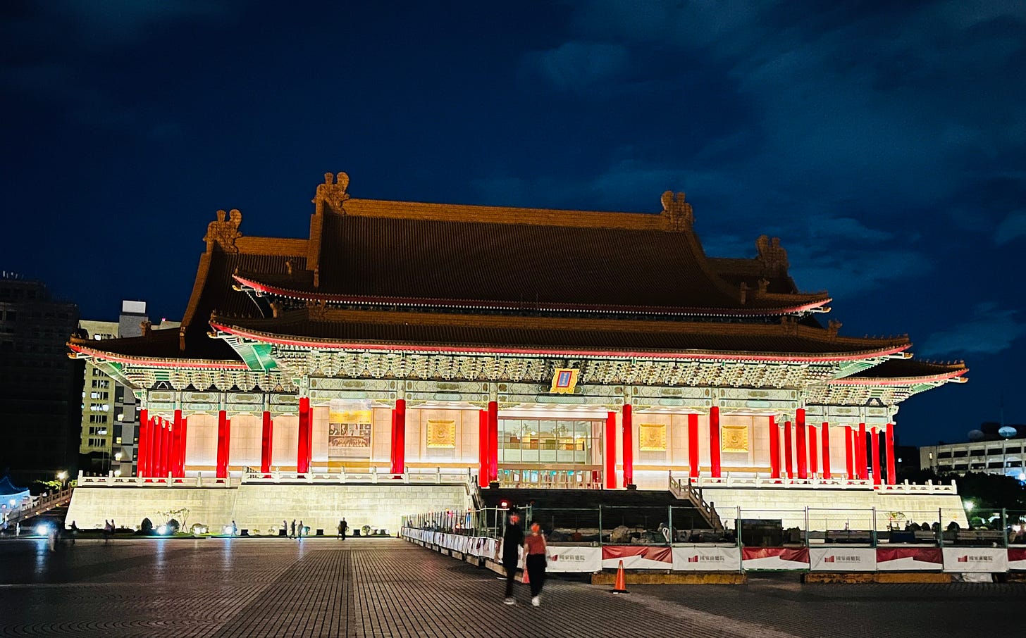 The exterior of Taiwan's National Concert Hall in Taipei at night. The building is illuminated to show off bright red columns on the entrance level underneath the upturned eaves of a yellow-tiled roof. In the square in front of the concert hall, a large area is fenced off for maintenance.