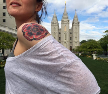 Karrie stands with her t-shirt draped over her shoulder to reveal the rose tattoo on her left shoulder. The photo cuts off so you cannot see her face, only her lips, slightly parted. In the distance behind her is the Salt Lake Temple.