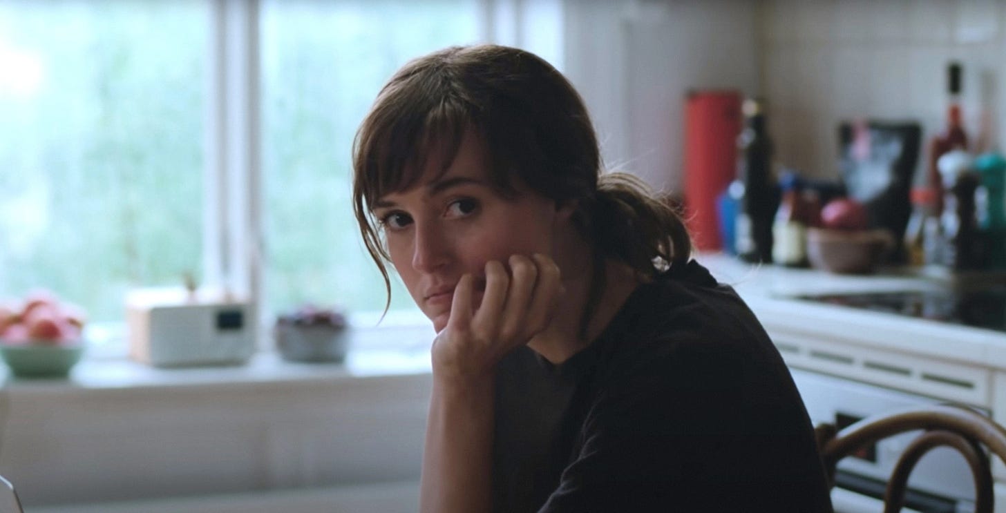 Movie still from The Worst Person in the World. A woman sits in a kitchen, hand on her chin.