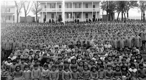 Black and white photograph featuring hundreds of young native children in front of a large white boarding house circa 1885. Black and white photograph featuring hundreds of young native children in front of a large white boarding house circa 1885.