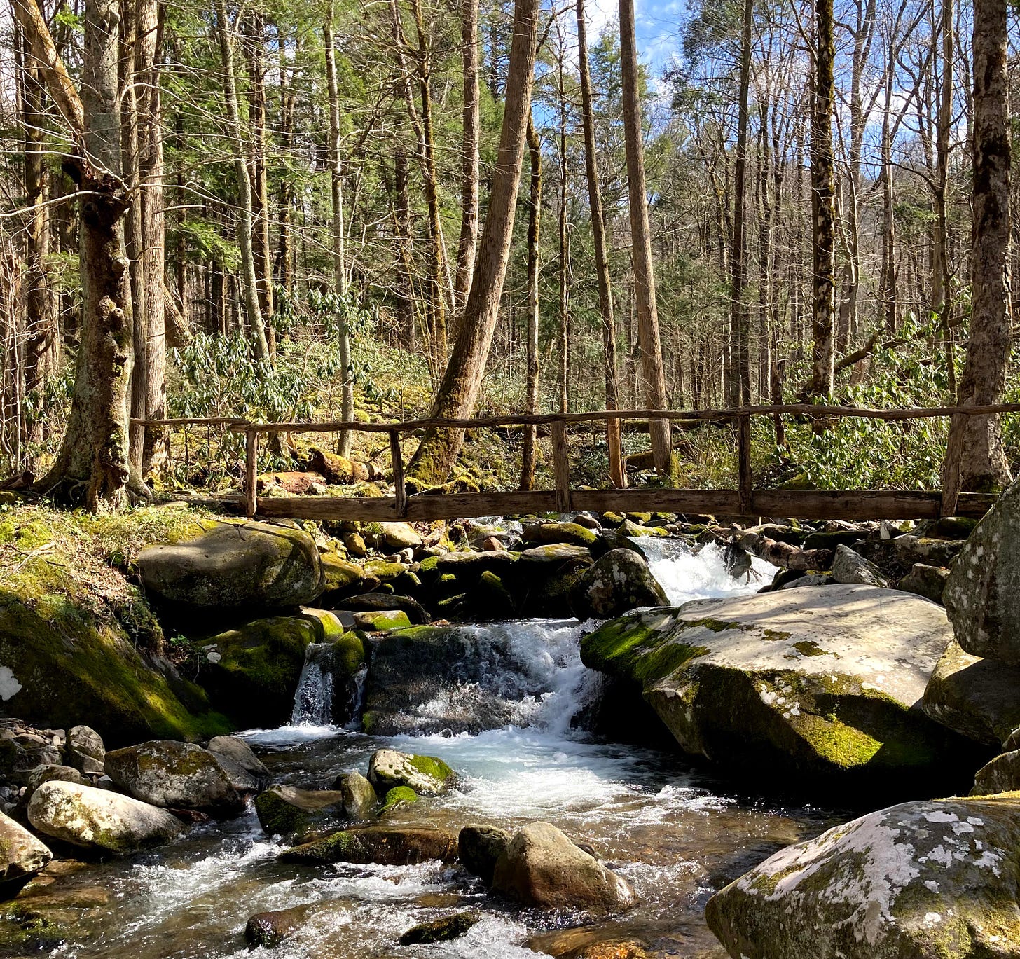 scenic view of a rough wooden bridge stretching over a creek running through granite rocks and bare trees, under a blue sky.