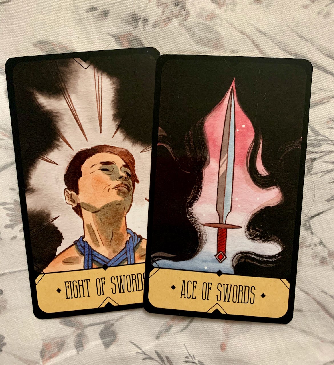 Eight of swords is a man’s face, eyes closed, surrounded by darkness. Eight sword points are poking out from the dark. The Ace of Swords is the same swirling darkness, but a single sword is shown clearly through it, on  a red and blue background. 