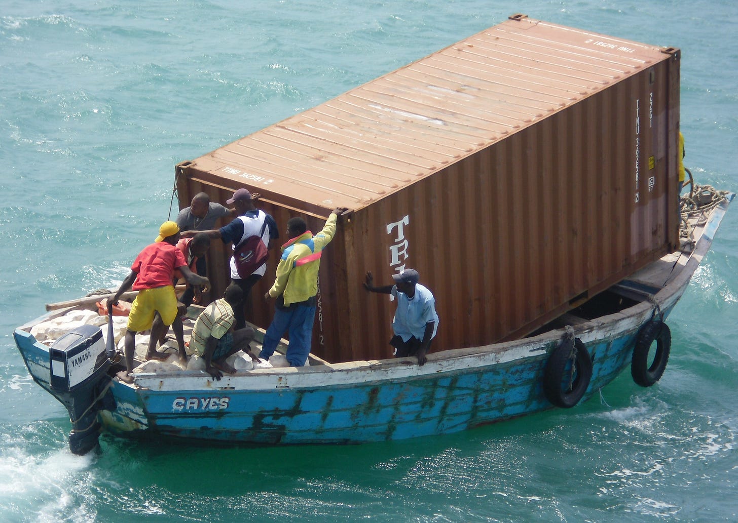 A single shipping container being transported on a small, ragged motorboat. A single shipping container being transported on a small, ragged motorboat.