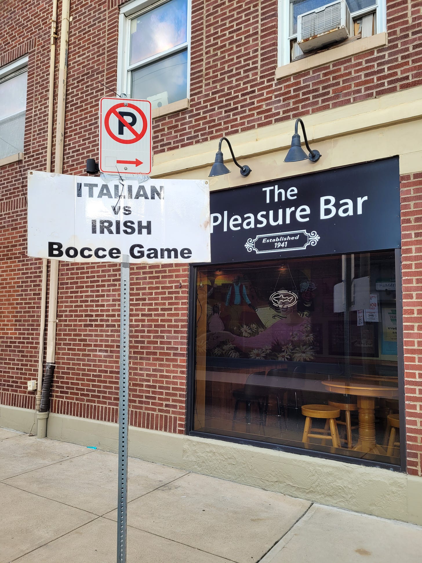 In the foreground: A sign reads "Italian vs Irish Bocce Game". In the background, a restaurant with a sign that reads "The Pleasure Bar"