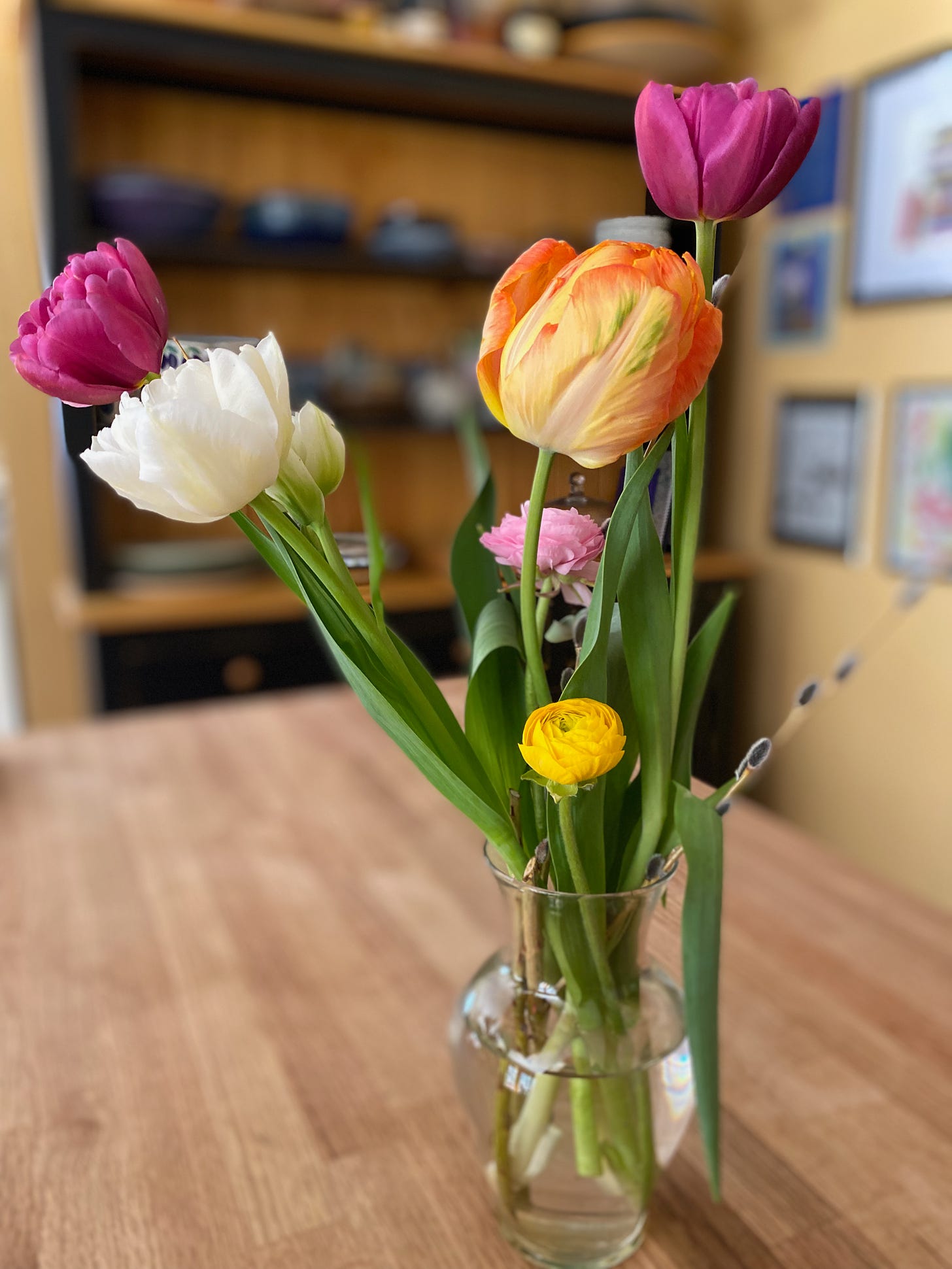 A glass vase of pink, orange, and white tulips, yellow ranunculus, and branches of pussy willow.