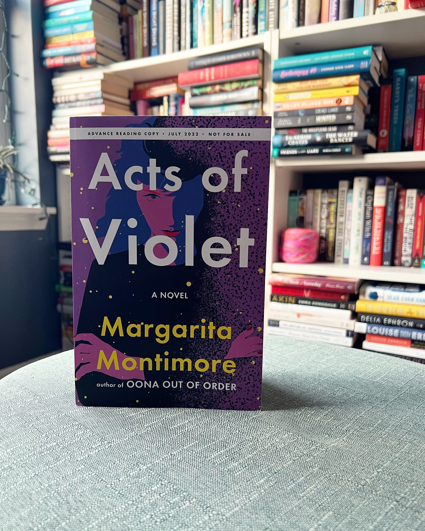 An ARC of Acts of Violet sitting on a cushion with lots of bookshelves behind it. 