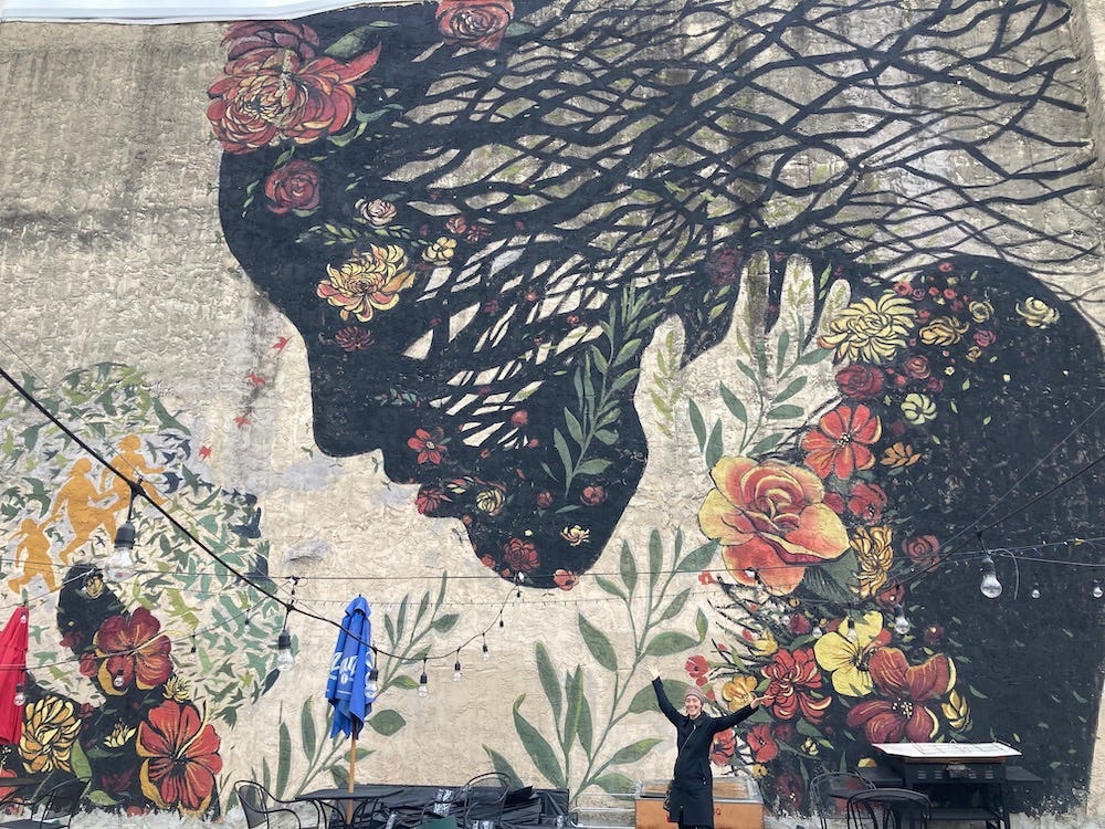Mural of two figures looking at each other, like parent and child. The larger figure is made from trees and flowers and roots, the smaller facemade from birds and people. I am standing at the bottom of the mural, barely visible in a black coat, arms outstreatched.
