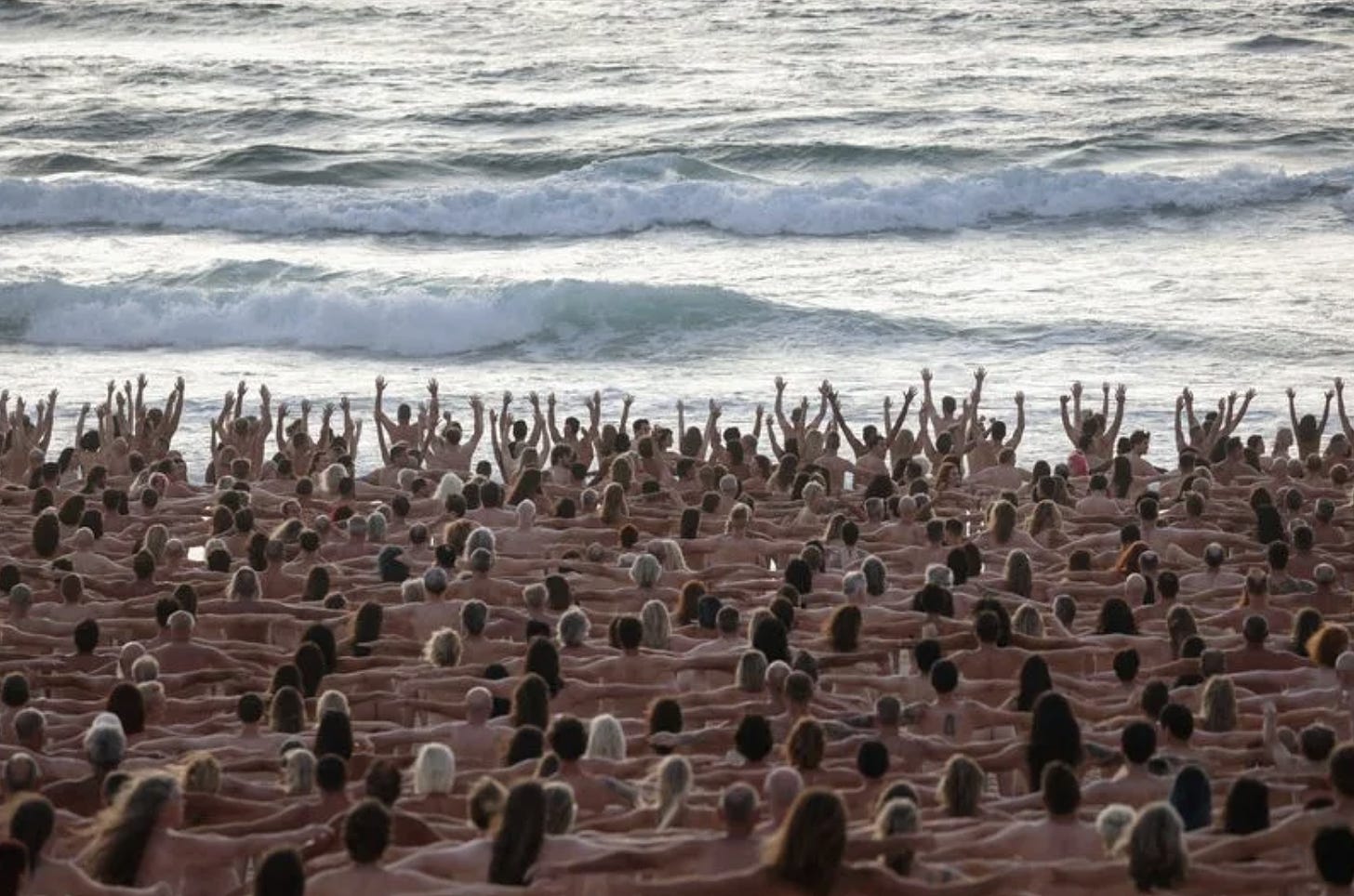 Hundreds of naked white people gathered and holding hands before an ocean shoreline. Some closest to the water have their hands raised.