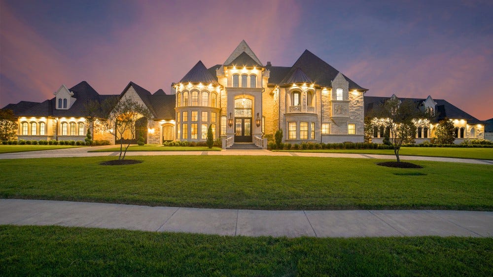 An obscenely large McMansion, faced with what appears to be rough fake stone, illuminated with all lights inside blazing yellow in the purple dusk. A wide swath of lawn extends from the circular drive to a sidewalk close to the camera.