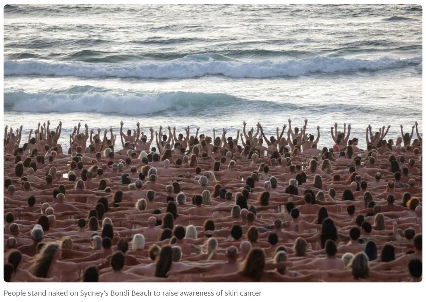 Hundreds of naked white people gathered and holding hands before an ocean shoreline. Some closest to the water have their hands raised. Caption at the bottom in black font on a white background reads: "People stand naked on Sydney's Bondi Beach to raise awareness of skin cancer."