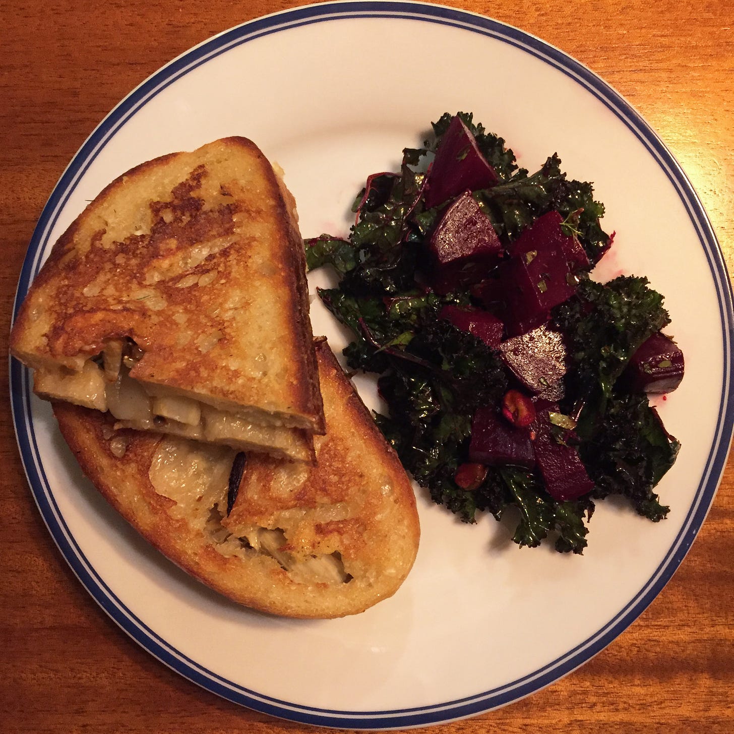 From above, a white dinner plate with two halves of a grilled cheese stacked on top of each other, on sourdough bread. Next to it is a dark red beet salad with pieces of kale and herbs visible throughout.