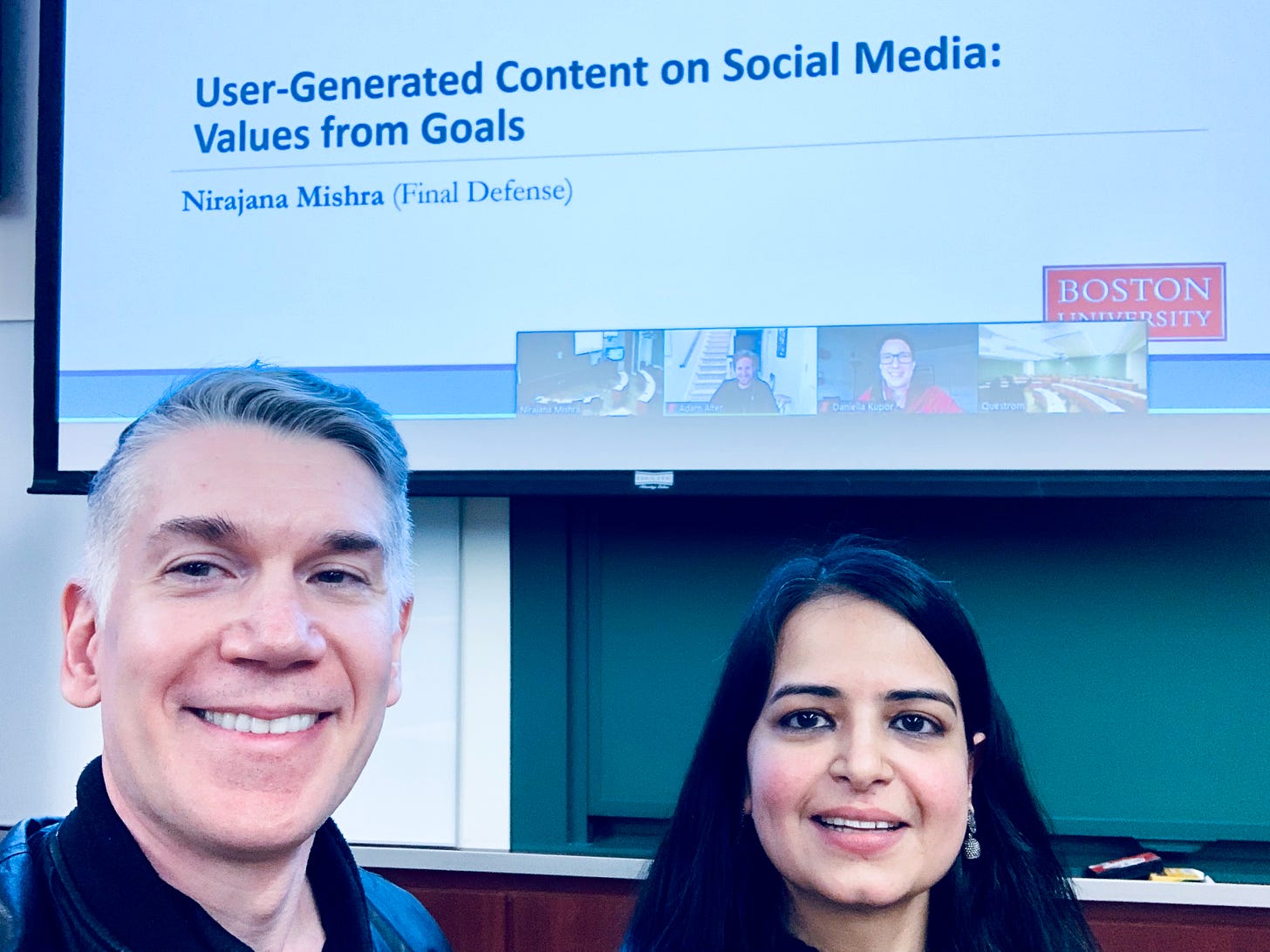 Dr. Carey Morewedge (left) and Dr. Nirajana Mishra (right) at her dissertation defense. A title slide for Dr. Mishra's presentation is in the background and reads "User-Generated Content on Social Media: Values from Goals."