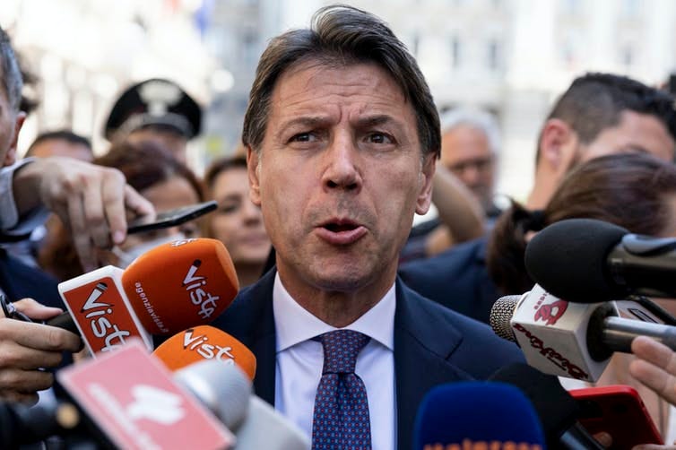 Close up of Giuseppe Conte's face, surrounded by microphones shoved in his face by members of the press
